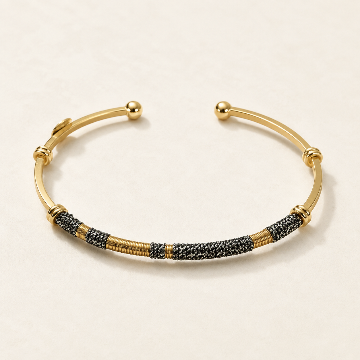 A gold-toned open cuff bracelet featuring textured black and gold woven segments along the lower curve, photographed on a clean off-white background.