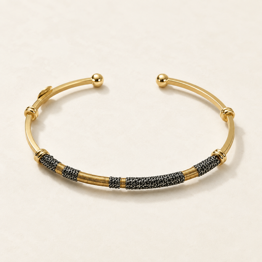 A gold-toned open cuff bracelet featuring textured black and gold woven segments along the lower curve, photographed on a clean off-white background.