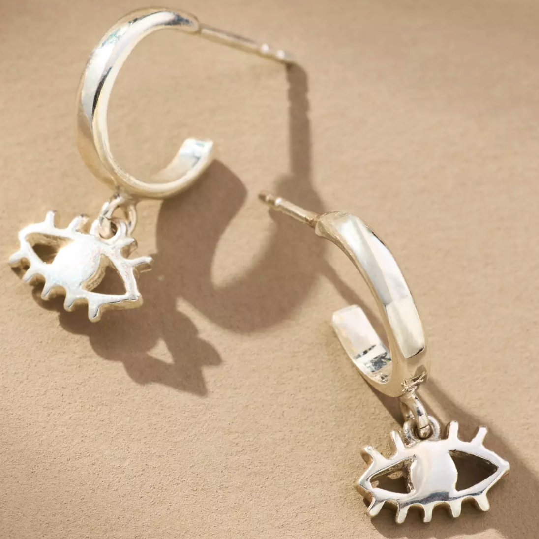 A pair of silver huggie hoop earrings with dangling eye charms, displayed on a textured beige surface with soft shadows.