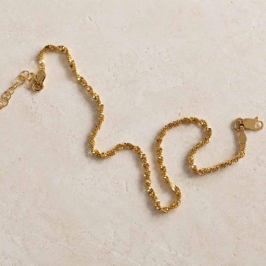 A gold chain necklace with a textured, rope-like link pattern, featuring a lobster clasp and an adjustable extension chain, displayed on a light beige stone surface.