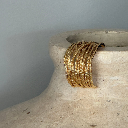 A wide, multi-strand gold bangle bracelet draped over the rim of a textured beige ceramic vase against a neutral gray background.