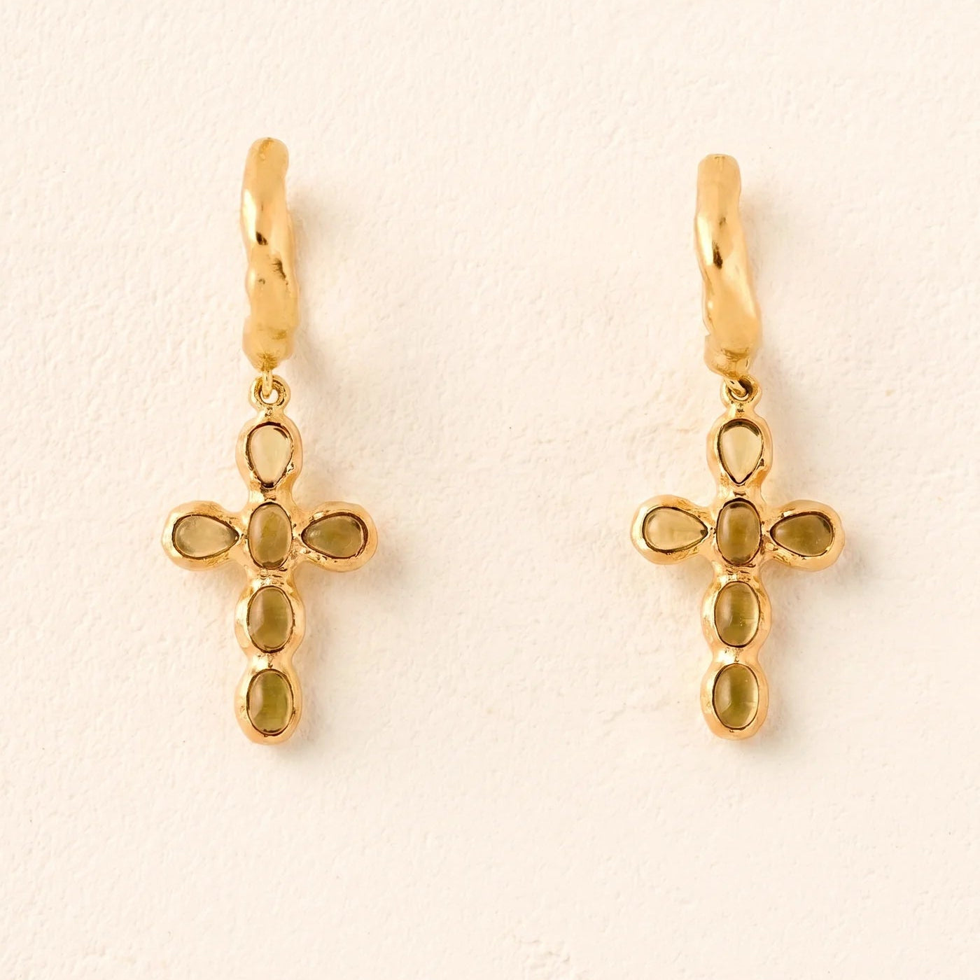 Gold cross-shaped earrings with green gemstones on a beige background
