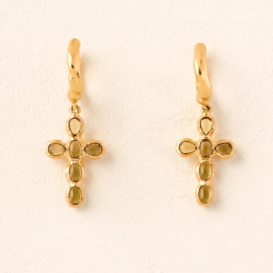 Gold cross-shaped earrings with green gemstones on a beige background