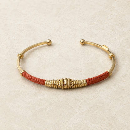 A gold-plated cuff bracelet featuring a central textured gold spool and rose clay braided cord on a light beige textured background.