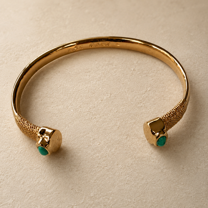A gold-toned open cuff bracelet with textured spiral ends and small green stone accents, photographed on a warm beige surface.