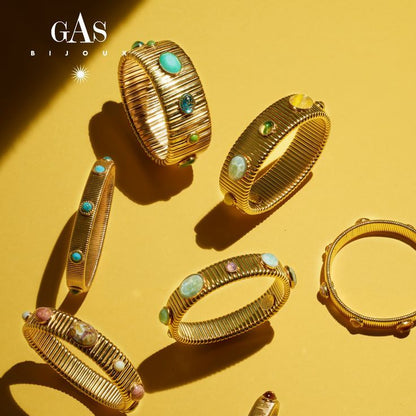 Six gold-toned bangle bracelets with textured ridges and colorful gemstone accents are arranged on a warm yellow background with dramatic shadows. The brand name GAS BIJOU is visible in the top left corner.