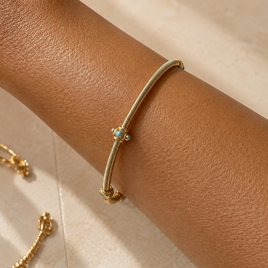 A close-up of a person's forearm wearing a slim gold bangle bracelet with a small turquoise and accent at the center, against a warm beige tiled background.