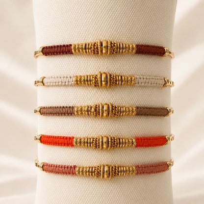 Five woven bracelet bands displayed on a cream fabric stand, each featuring textured gold-toned beadwork and spacer details. The bands are in brown, light grey, taupe, orange and rose clay.